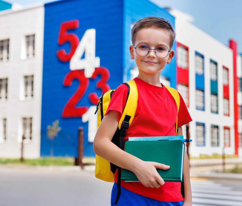 Boy with glasses and a backpack holds a book in his hands against the background of a modern school.
