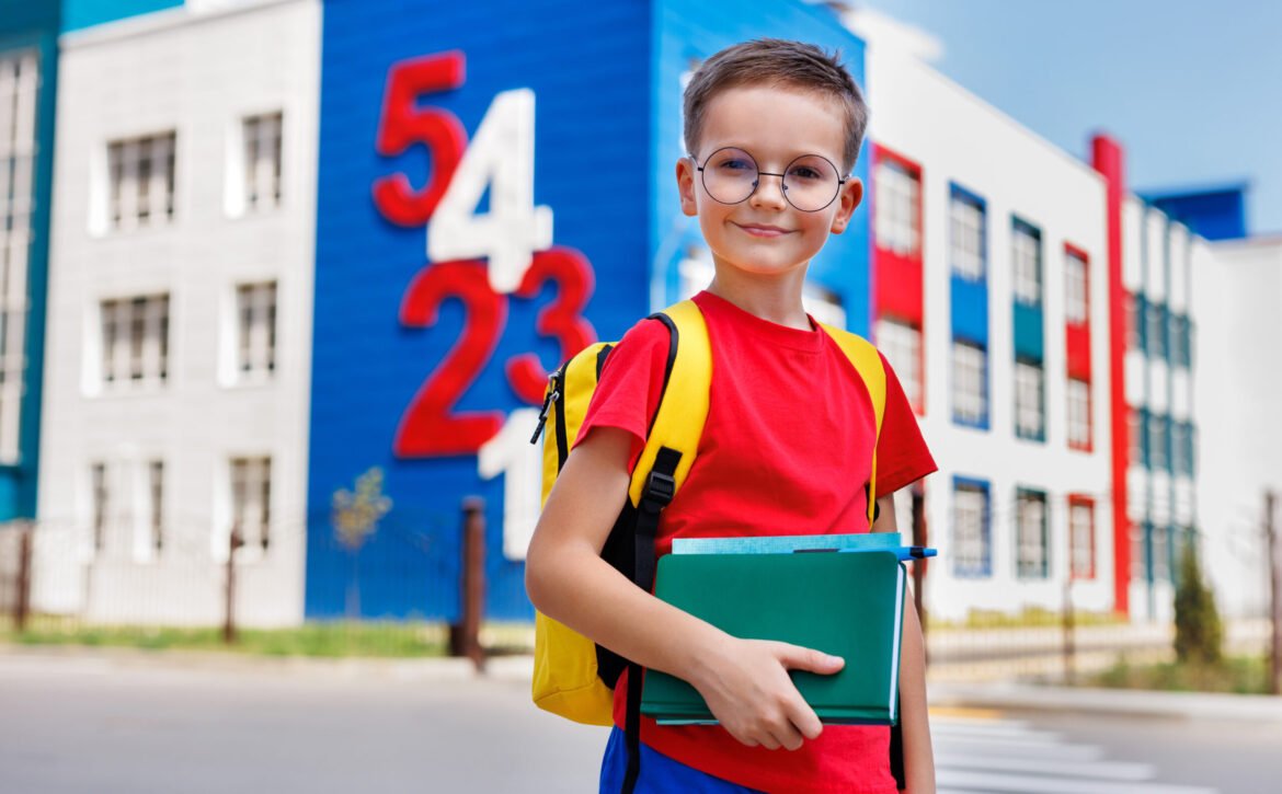 Boy with glasses and a backpack holds a book in his hands against the background of a modern school.